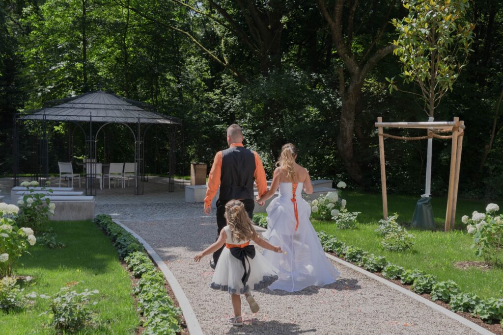 Hochzeitspaar schreitet zum Pavillon im Edla Park. Die tochter der beiden läuft in einem weißen Kleid hinterher.