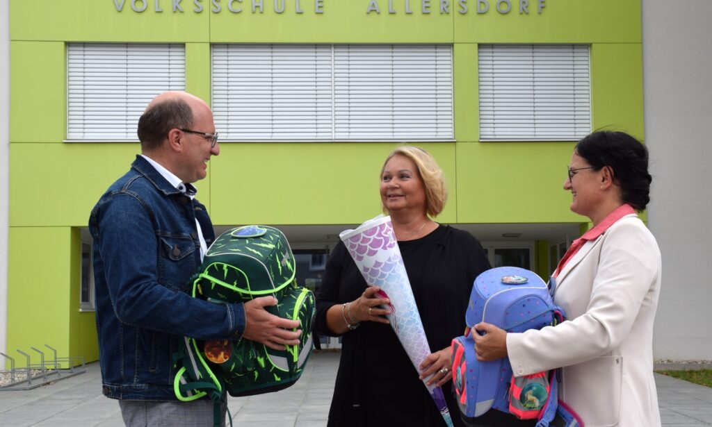 Schulausschuss-Obmann Reinhard Aigner, MA; Bildungsstadträtin Doris Koch, MSc und Bildungsgemeinderätin Helga Seibezeder stehen vor der Volksschule Allersdorf mit Schultaschen und Schultüte in der Hand.